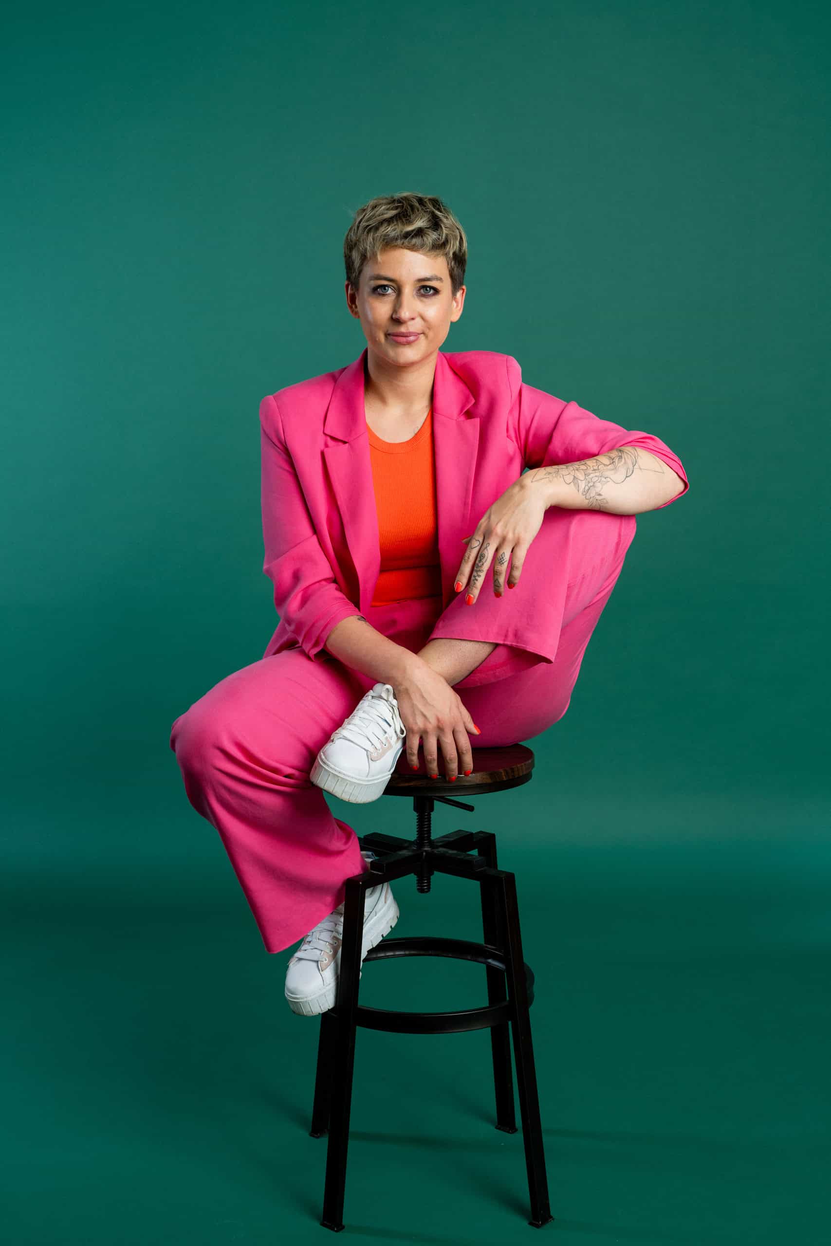 boxquadrat-paula-sidorczuk-2025. Person im pinken Anzug sitzt auf Hocker vor grünem Hintergrund - boxquadrat