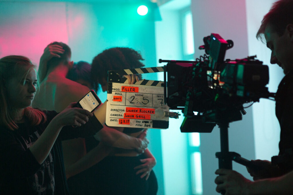 Eine Filmkamera die eine Filmklappe filmt. Im Hintergrund drei Tänzer in einem grün, rosa Raum