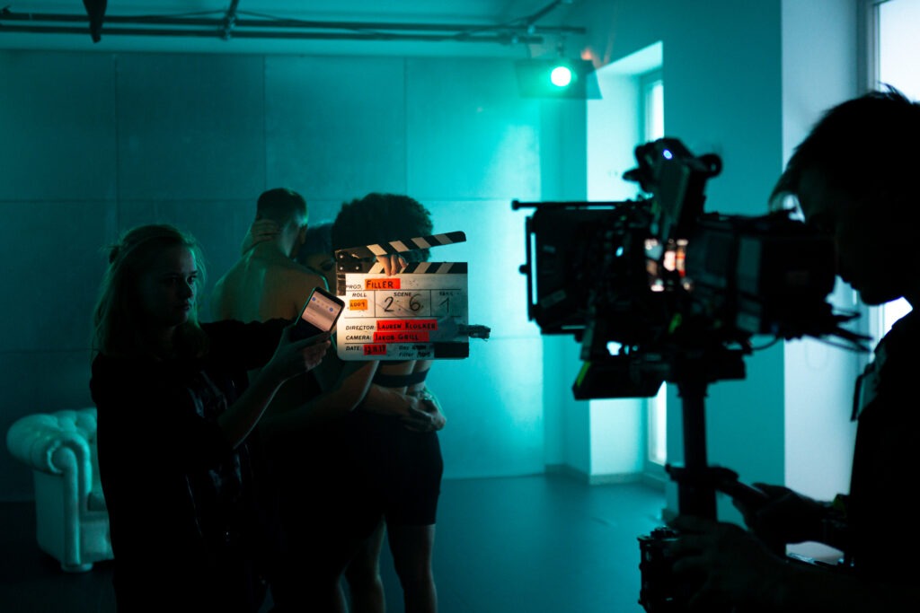 Eine Filmkamera die eine Filmklappe filmt. Im Hintergrund drei Tänzer in einem grün, blauen Raum