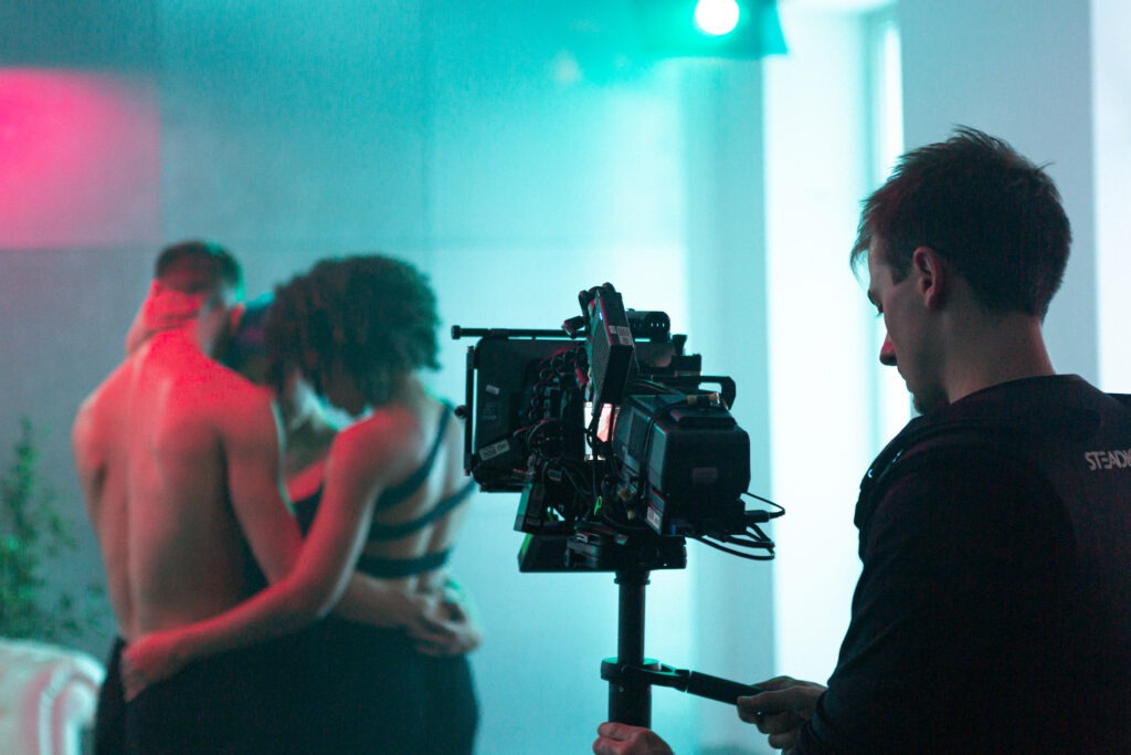 Eine Filmkamera. Im Hintergrund drei Tänzer in einem blau, rosa Raum