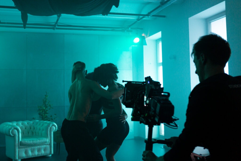 Eine Filmkamera. Im Hintergrund drei Tänzer in einem grün, rosa Raum