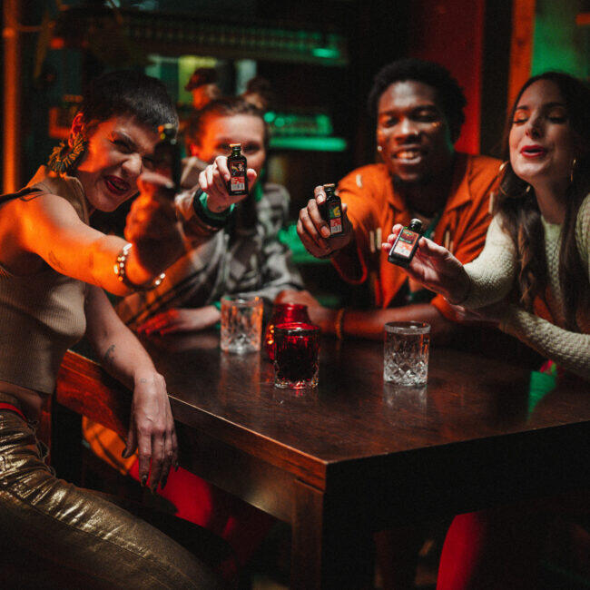 Fünf junge Erwachsene sitzen an einem Holztisch in einer Bar mit stimmungsvoller, bunter Beleuchtung. Sie halten Miniaturflaschen eines Kräuterlikörs von Jägermeister in die Kamera und lächeln oder lachen. Auf dem Tisch stehen mehrere Gläser mit klaren und roten Getränken. Die Szene wirkt lebendig und gesellig, mit Party- oder Feieratmosphäre.