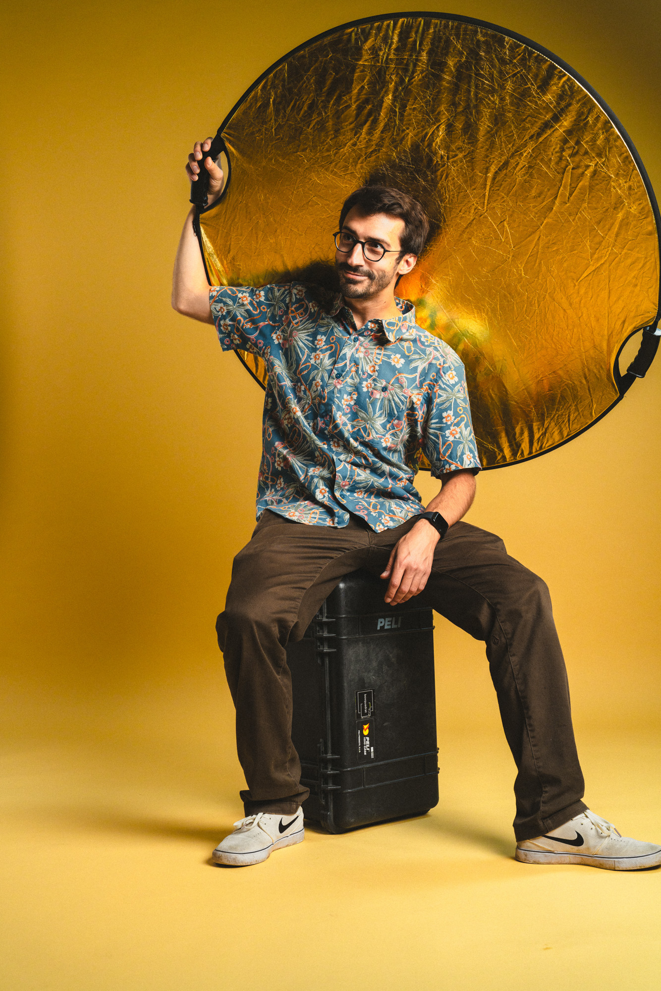 Ganzkörper Foto von Jakob Grill, sitzend auf einem Pelicase mit einem goldenen Faltreflektor in der Hand, im boxquadrat Fotostudio 1160 Wien