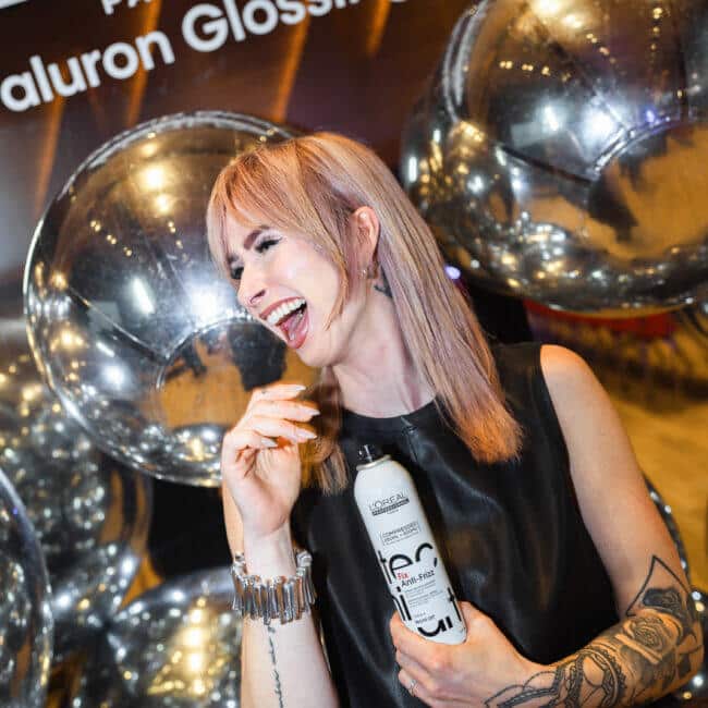Frau mit rosa-blondem Haar posiert vor dem L'Oréal Hyaluron Glossing Backdrop mit silbernen Ballons