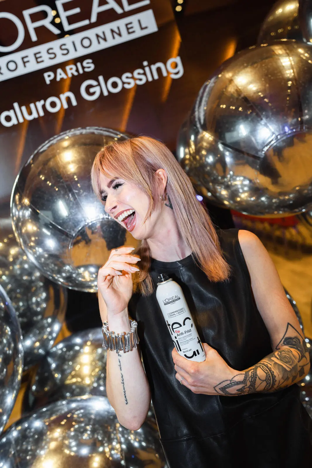 Frau mit rosa-blondem Haar posiert vor dem L'Oréal Hyaluron Glossing Backdrop mit silbernen Ballons